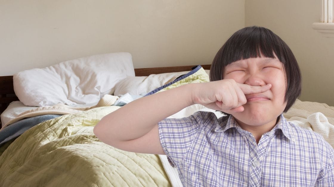 A young asian girl sneezing in front of a bed.