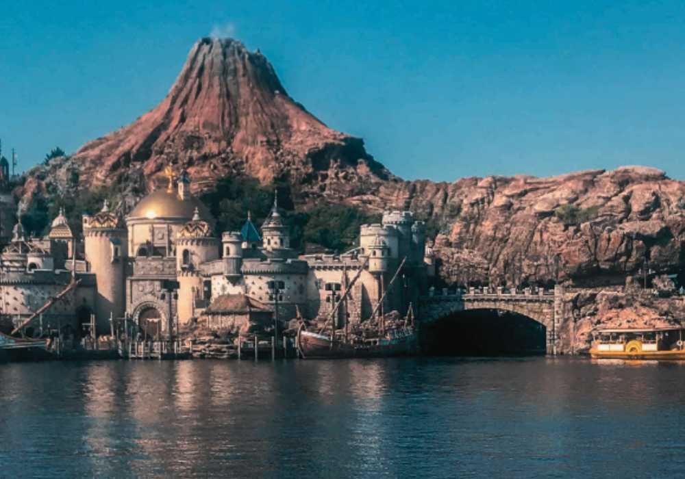 A picturesque view of a theme park with a stylized mountain backdrop and european-inspired architecture by a calm river.