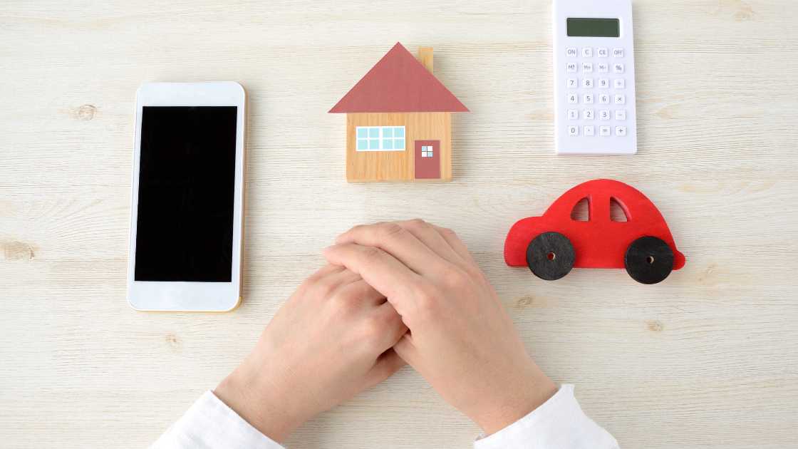 A person seated at a table with a smartphone, calculator, small house model, and car toy, symbolizing personal finance planning related to homeownership and transportation.
