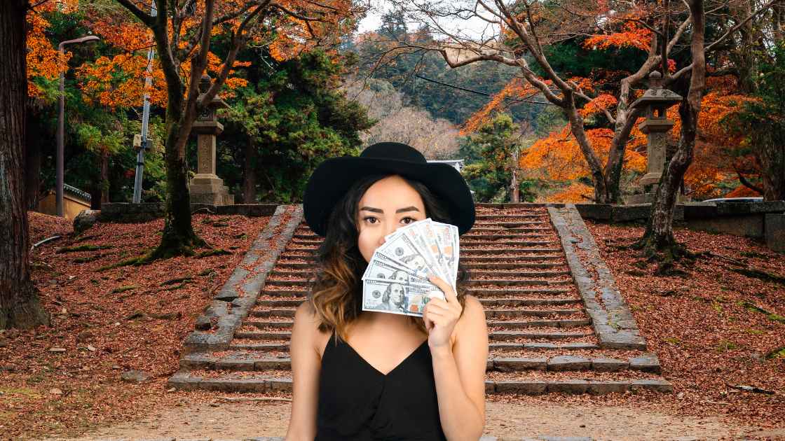 A woman in a black hat and top covers half her face with money, standing before a staircase surrounded by autumn trees.