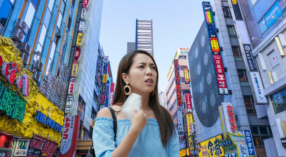 A woman stands in a busy city street with tall buildings and numerous colorful signs, holding a tissue to her face with a concerned expression.