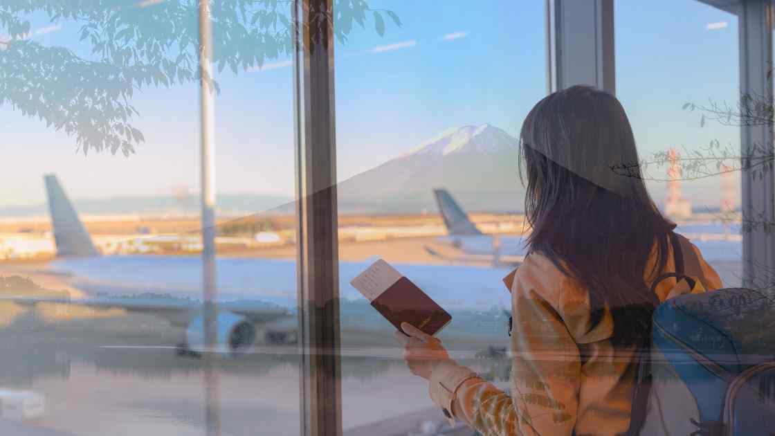 Woman holding a passport in an airport lounge, looking out a window at mount fuji in the distance, with airplane tails visible.