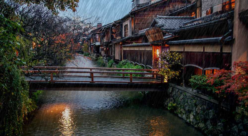 Rain falls on a narrow street with traditional wooden buildings, a small bridge over a stream, and colorful autumn foliage.