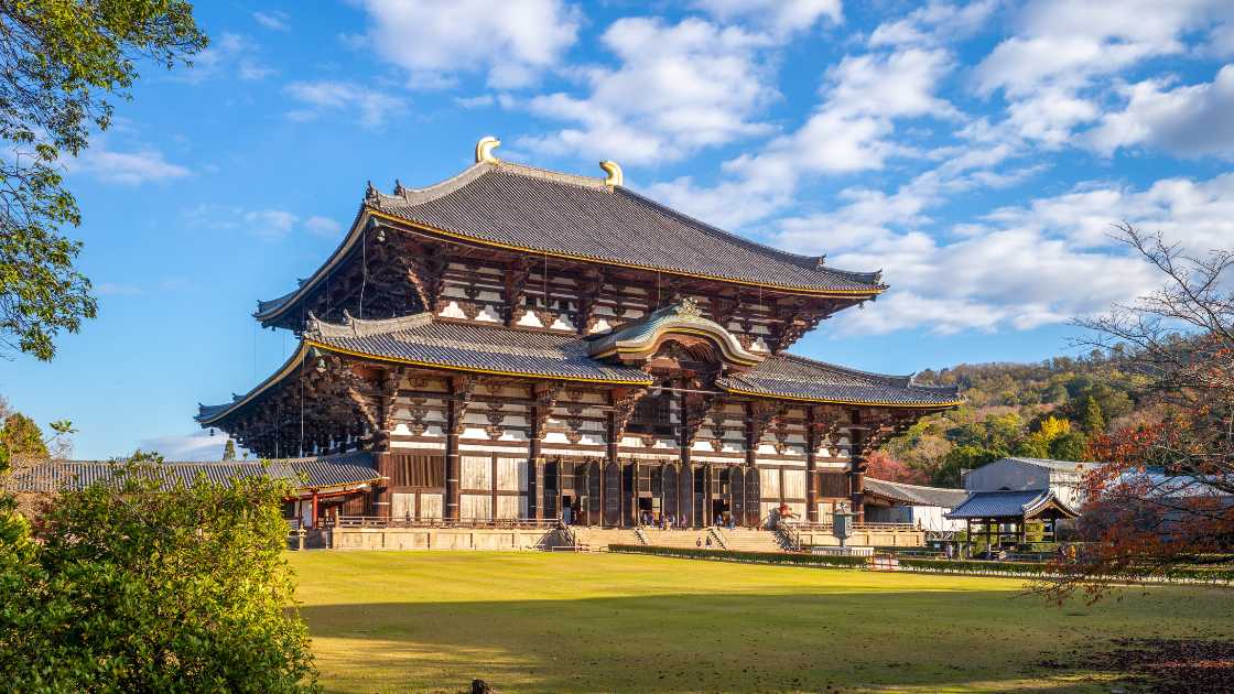 Large traditional japanese temple with sweeping roofs, situated in a scenic, lush park under a clear blue sky.