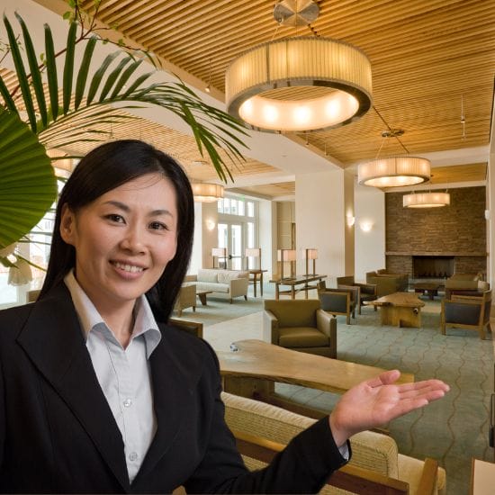 A person in business attire gestures towards a spacious, well-lit room with modern chandeliers, wooden ceiling, armchairs, sofas, and a fireplace in the background.