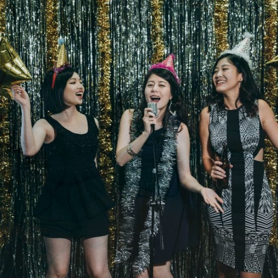 Three women, wearing festive hats and party attire, sing into a microphone in front of a sparkling, tinsel backdrop.