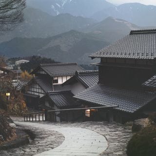 A peaceful, traditional Japanese village at dusk with tiled-roof houses lining a curved stone path, set against a backdrop of misty mountains.