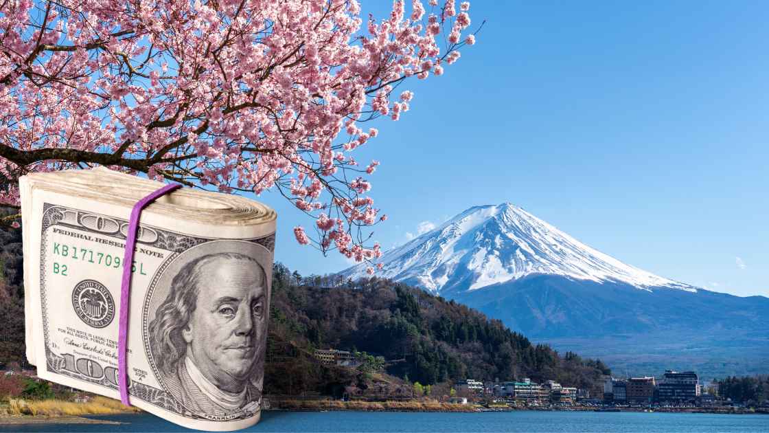 A rolled-up dollar bill with Benjamin Franklin's face, secured with a pink ribbon, beside a blooming cherry blossom tree in Japan, with Mount Fuji and a clear blue sky in the background.