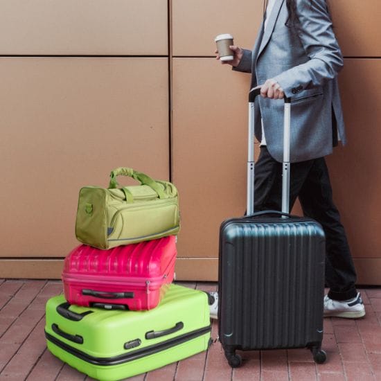 A person standing next to stacked luggage, holding a rolling suitcase and a coffee cup. The luggage includes bright pink, green, and black bags.