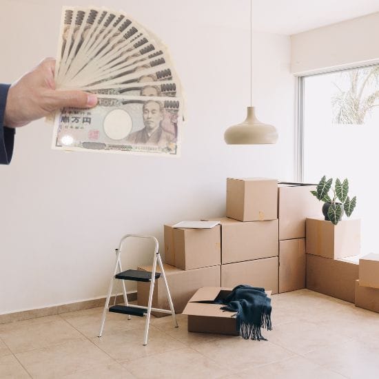A hand holding Japanese yen banknotes. In the background, there are several stacked cardboard boxes, a step ladder, and a potted plant in a bright, empty room.