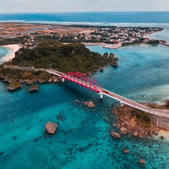 Aerial view of a red arched bridge connecting two land masses over clear turquoise waters, with a small town, lush greenery, and coastline in the background.