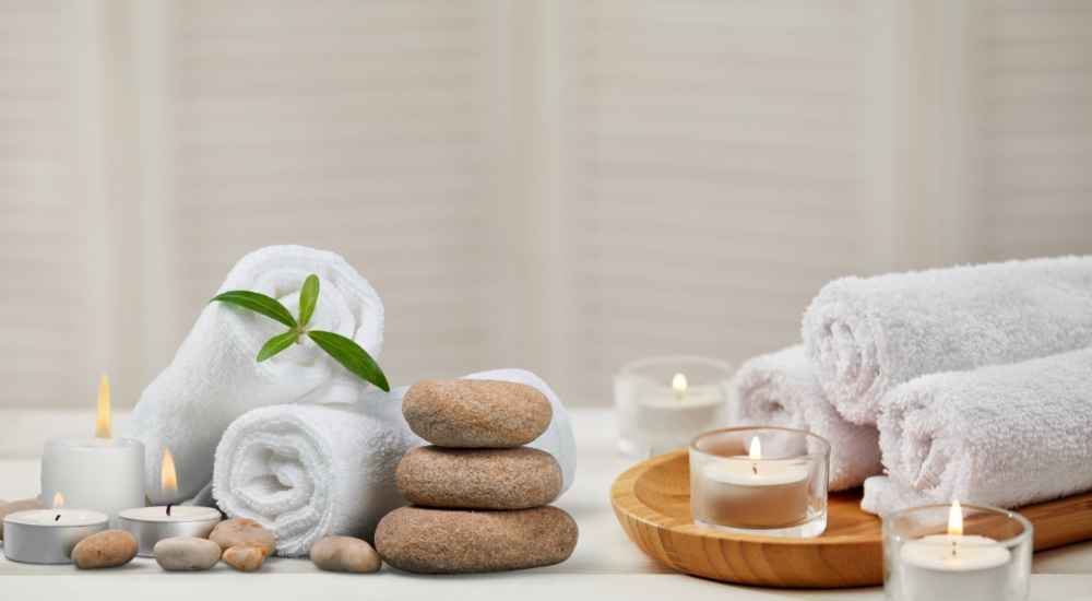 Rolled white towels, stacked stones, small candles, and a wooden bowl on a table create a relaxing spa setting.