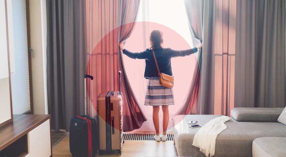 A person stands in a hotel room, pulling back curtains to look out the window. Luggage is beside them and a smartphone is on a nearby couch.