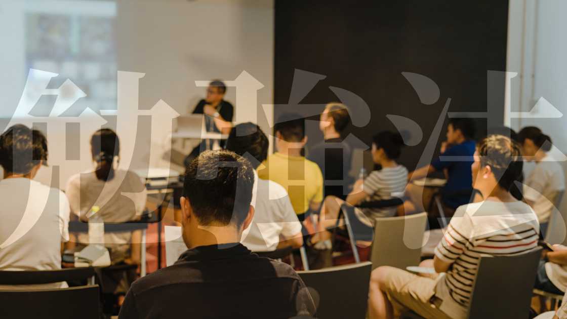 A group of adults seated, listening to a speaker at the front of a room during a seminar on "Why Choose Japan for Study: Unveiling the Benefits for International Students.