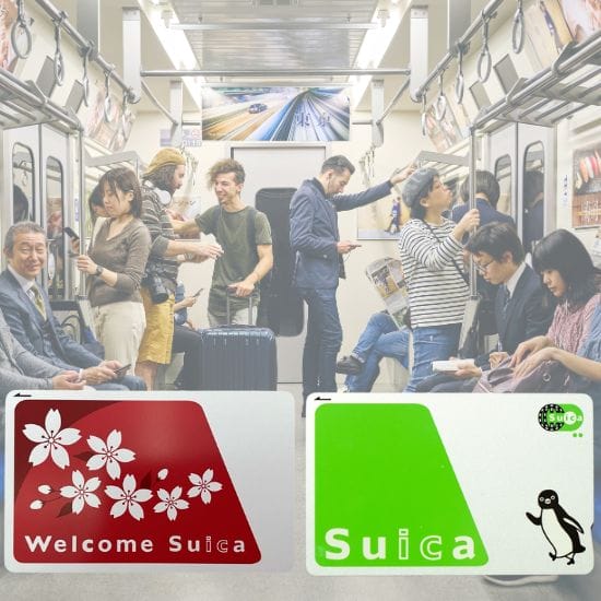 Passengers ride on a relatively crowded train. Some are standing and engaged in various activities while others are seated, looking at their phones or resting. Two Suica cards are shown in the foreground.