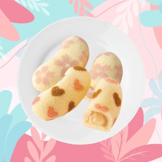 A white plate with four colorful, bean-shaped sponge cakes, one of which is bitten to reveal a creamy filling, rests against a vibrant, floral and abstract patterned background.