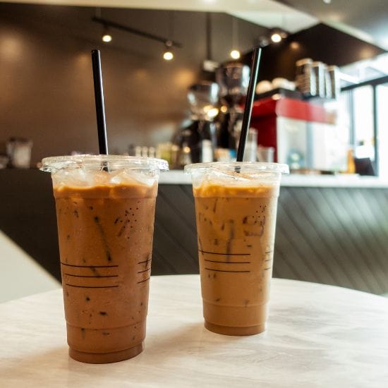 Two cold coffee drinks with black straws on a round table in a modern café setting.