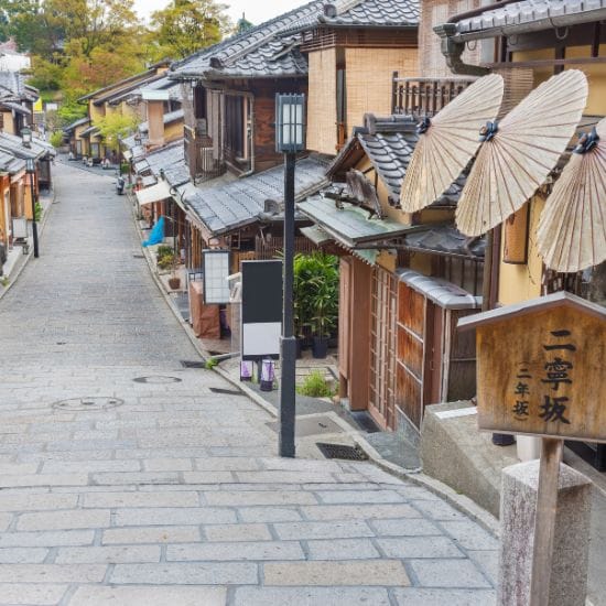 A narrow, sloped stone-paved street lined with traditional wooden buildings and parasols. Japanese signage is visible, and trees are in the background.