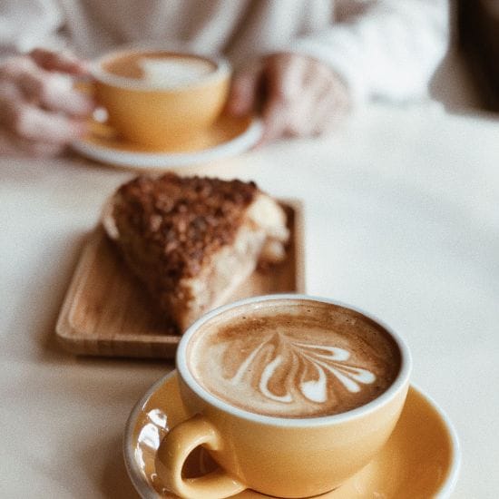 Two yellow cups of latte with latte art sit on a white table. A piece of pie on a wooden plate is in the background, with a person holding one of the cups.