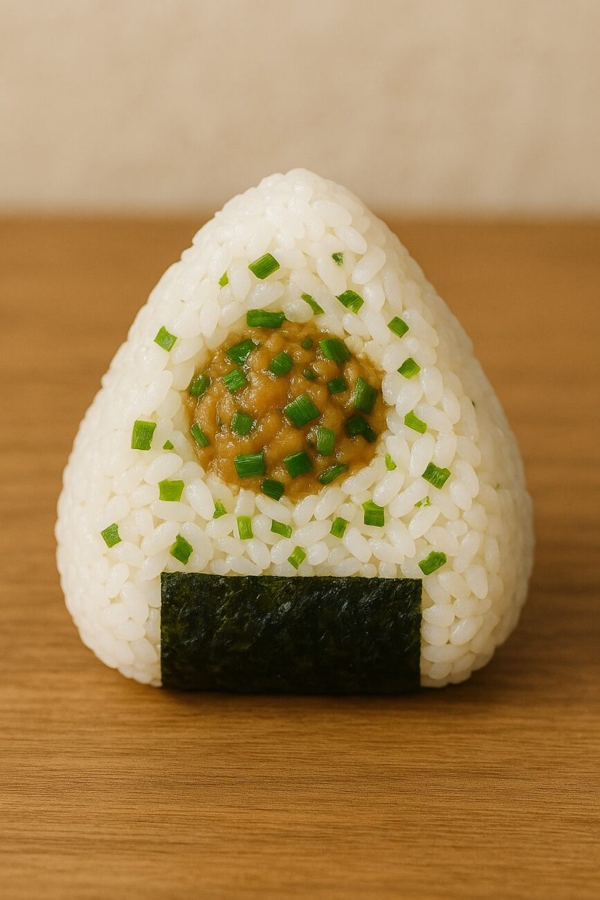 A triangular rice ball on a wooden surface, filled with chopped scallions and wrapped with a strip of nori.