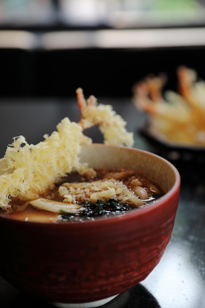 A black bowl of miso soup with green onions sits on a wooden tray next to a white spoon. The background features the text "japanwhisper.com".