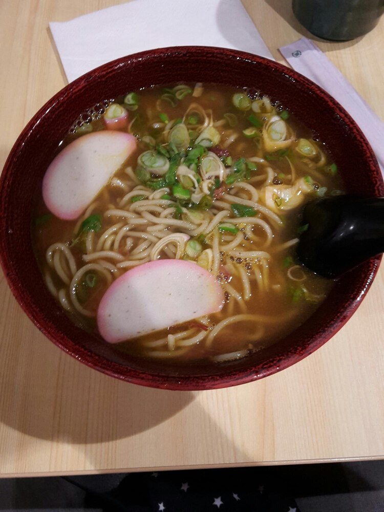 A bowl of udon noodle soup with seaweed, green onions, and broth on a wooden table, with chopsticks and a spoon in the background.