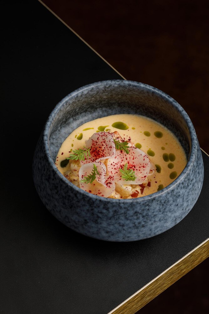 A bowl of soup with sliced meat, tofu, vegetables, and green onions, placed on a wooden tray near a window.