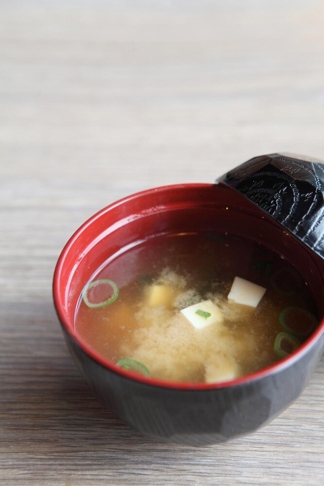 A bowl of miso soup with tofu cubes and seaweed, served on a wooden coaster with a spoon inside the bowl.