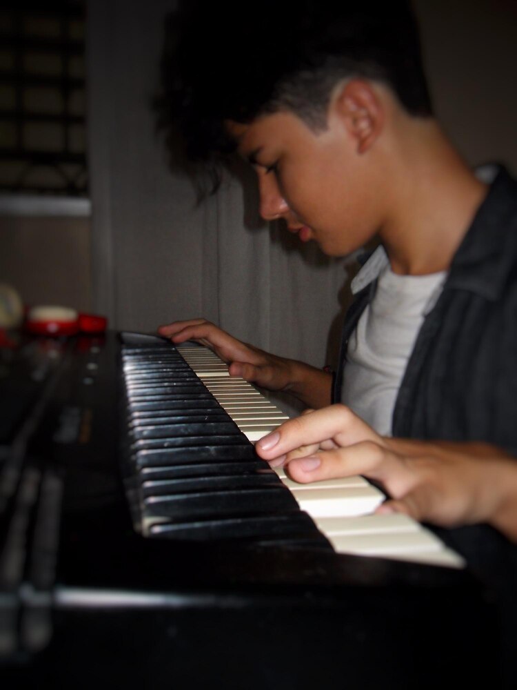 A person in a beige suit plays the piano next to a basket filled with sheet music, with flowers and sunlight streaming through sheer curtains in the background.
