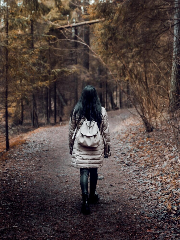 A person with long hair and a brown jacket walks alone on a sunlit forest path, surrounded by tall trees and green foliage.