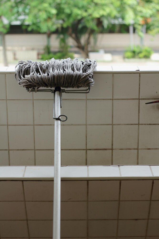 A blue cleaning brush is scrubbing a glass shower door with water droplets, with white tiled walls in the background.
